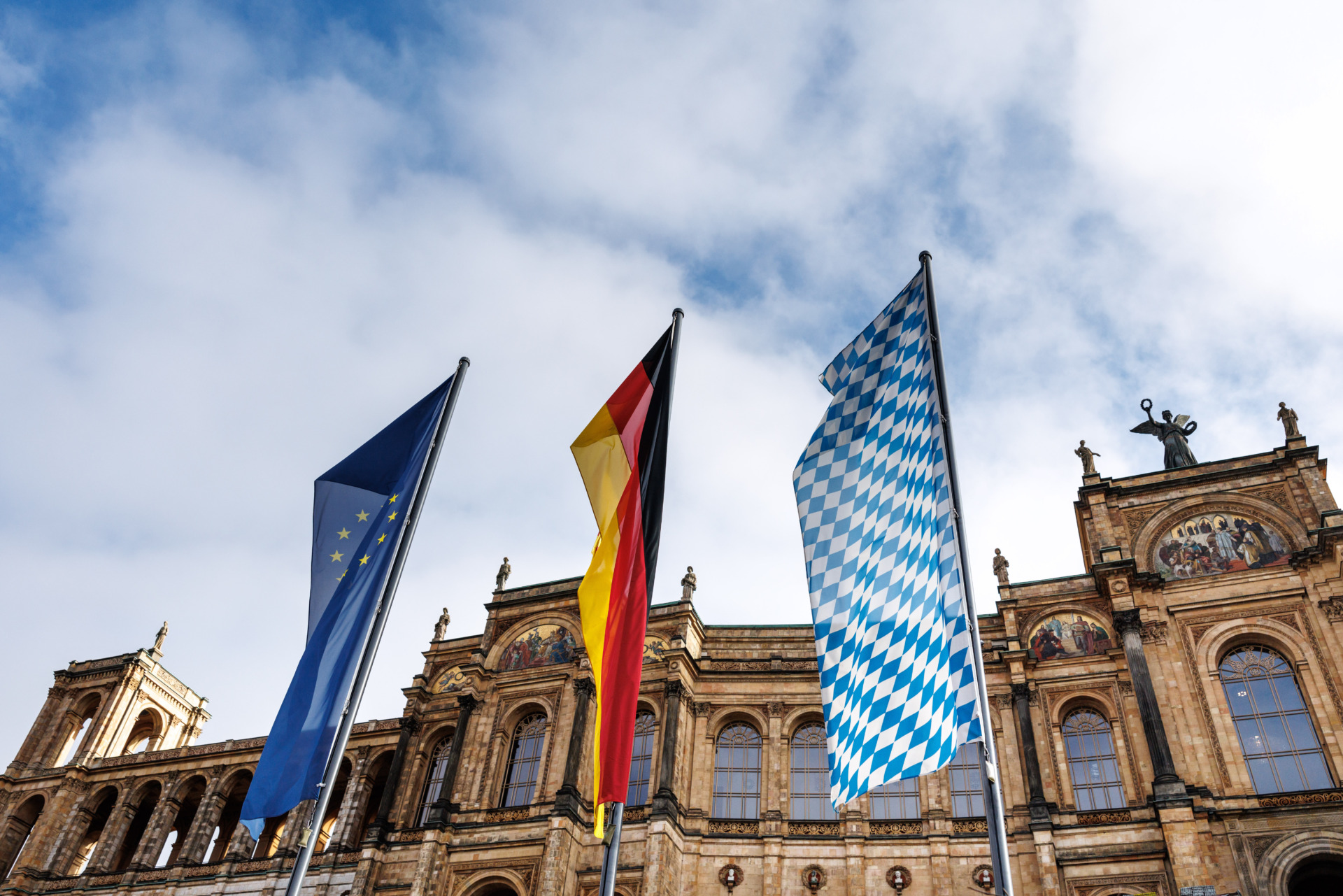 Der Bayerische Landtag in München ist abgebildet. Davor stehen drei große Flaggen: Europa, Deutschland und Bayern. Der Landtag beschäftigt sich jetzt mir dem True-Crime-Boom.