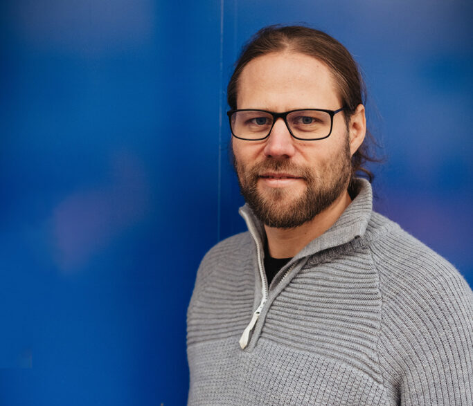 Ein Foto zeigt einen Mann mit Brille, der vor einer blauen Wand steht.