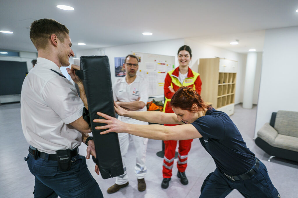 In einem Raum stehen vier Personen. Eine Frau drückt mit beiden Händen gegen eine Gummimatte, die ihr ein Mann entgegenhält.