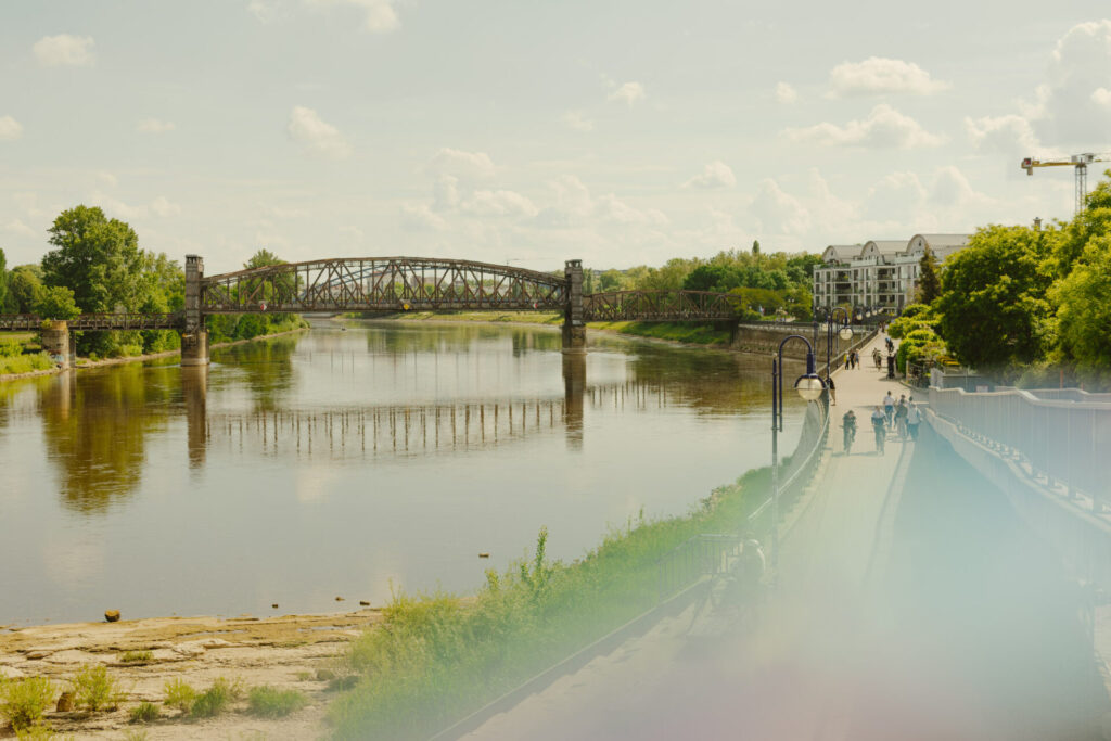 Menschen spazieren an der Elbe in Magdeburg entlang. Über der Elbe erstreckt sich die ehemalige Eisenbahnbrücke „Hubbrücke“.