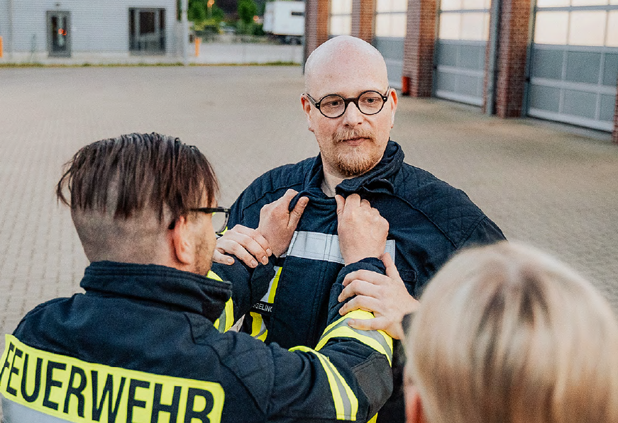 Eine Person in Feuerwehrkleidung hält eine andere Person bei einer Übung am Kragen fest. Die hintere Person schaut zu einer anderen Person, die im Vordergrund des Fotos von hinten zu sehen ist.