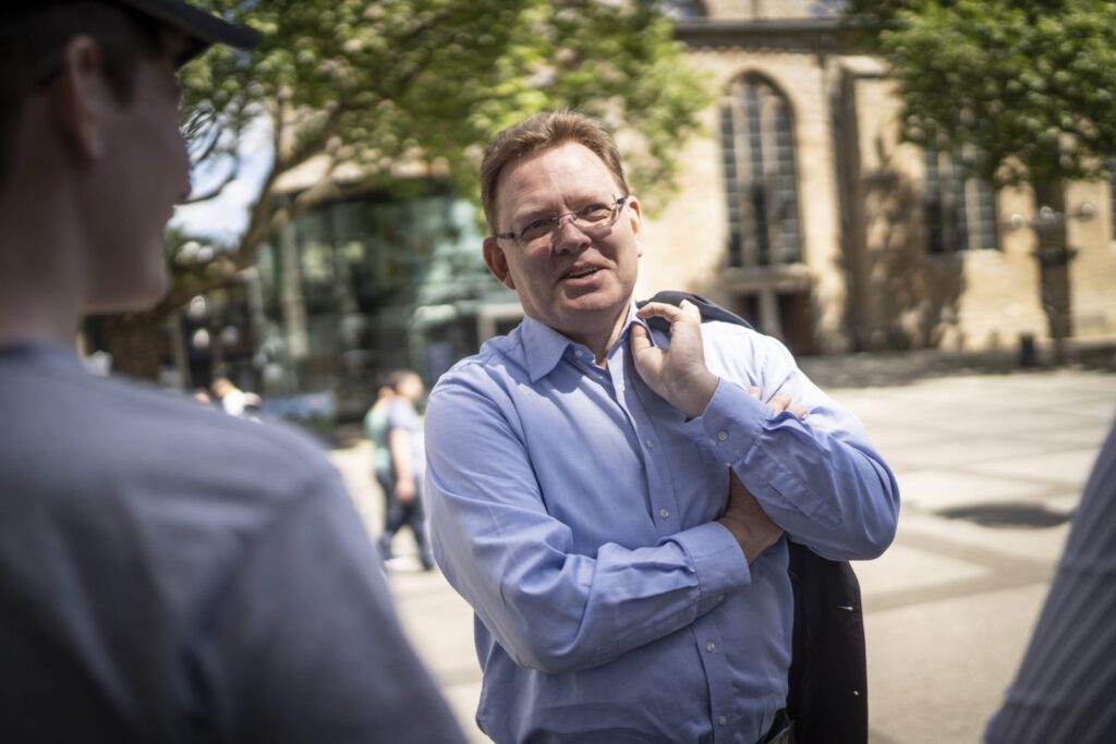 Ein Mann mit Brille hält ein Sakko über der Schulter.