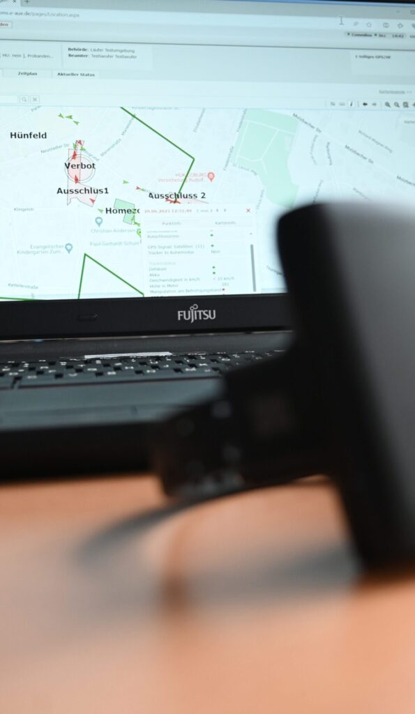 Elektronische Fußfessel liegt auf einem Tisch. Davor steht ein Laptop und auf dem Bildschirm sieht man geografische Angaben.