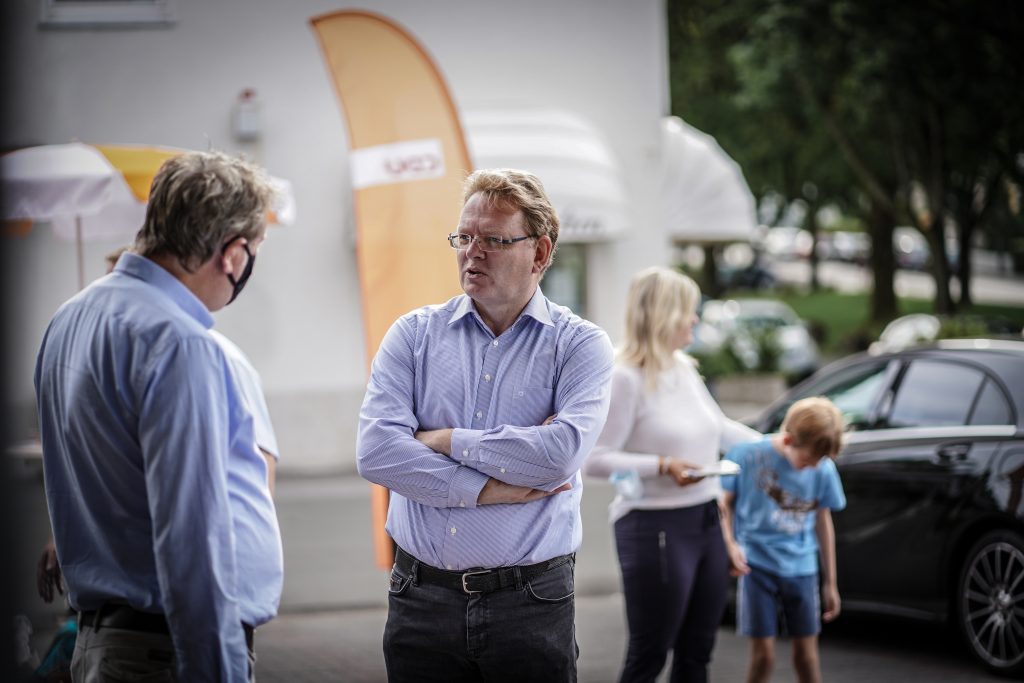 Ein Mann mit verschränkten Armen unterhält sich mit einer Person, die eine medizinische Maske trägt. Im Hintergrund stehen eine Frau und ein Kind